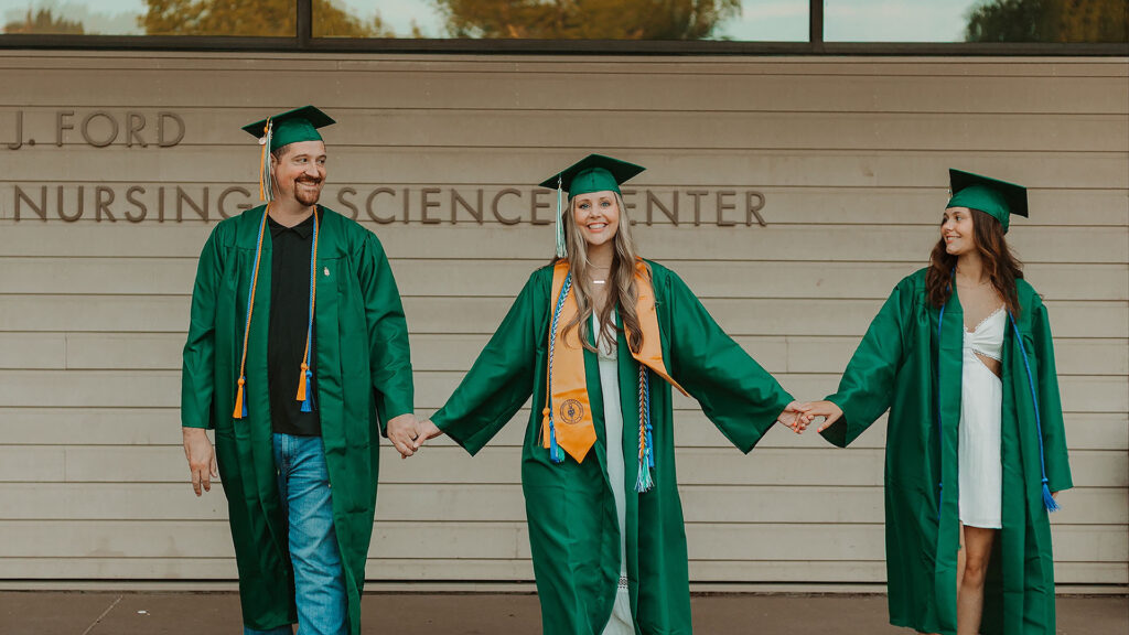 Three family members in green graduation caps and gowns hold hands outside the Ford Nursing Science Center, celebrating their UCC nursing graduation together.