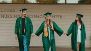 Three family members in green graduation caps and gowns hold hands outside the Ford Nursing Science Center, celebrating their UCC nursing graduation together.