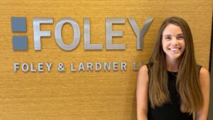 Sydney Miele standing and smiling in front of a Foley & Lardner LLP office sign, where she works as a legal assistant after graduating from UCC's Paralegal program.