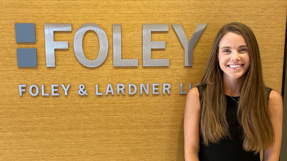 Sydney Miele standing and smiling in front of a Foley & Lardner LLP office sign, where she works as a legal assistant after graduating from UCC's Paralegal program.